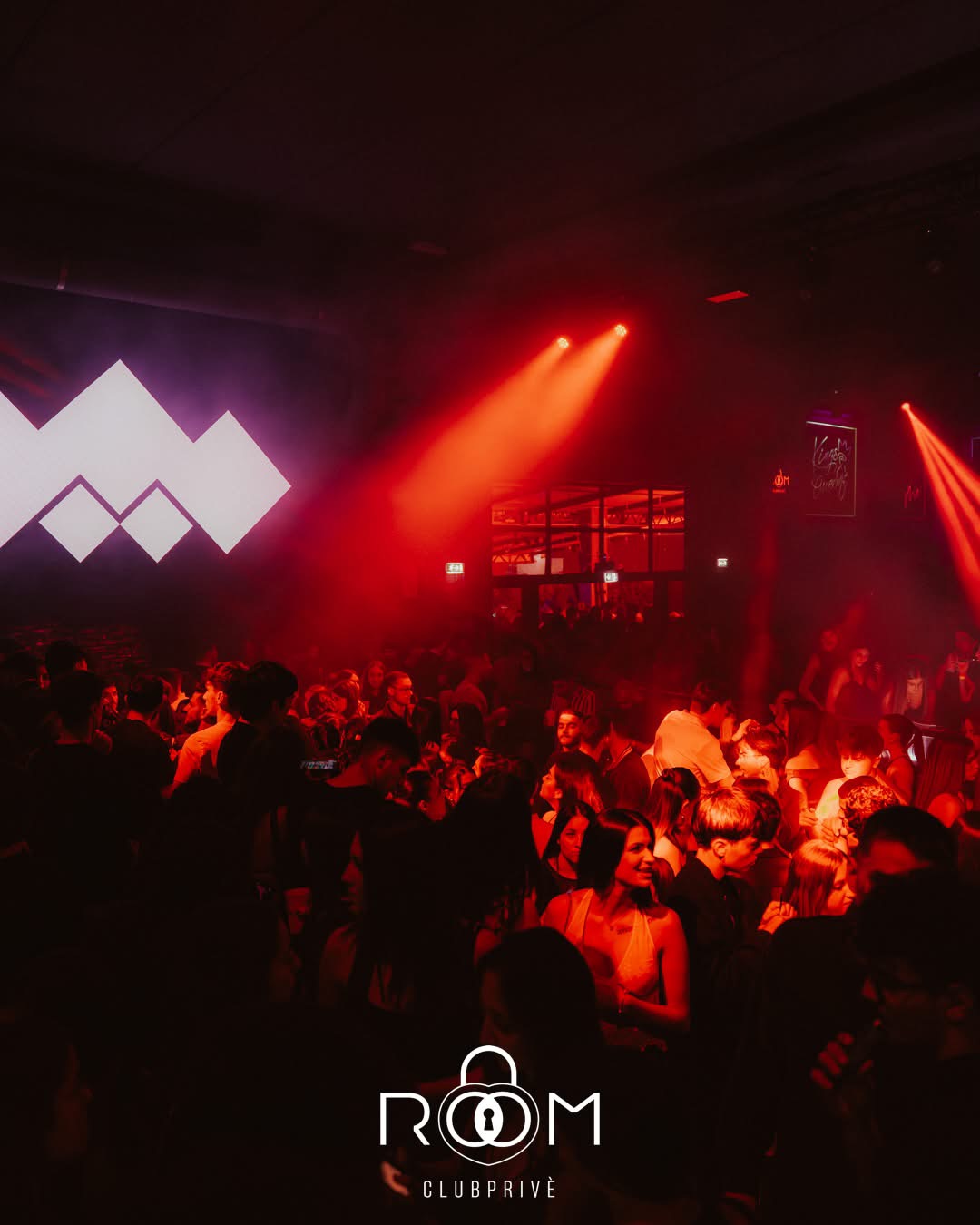 Il dancefloor del ROOM sotto i fasci di luce rossi, con un grande logo luminoso a rombi sulla parete di sinistra.