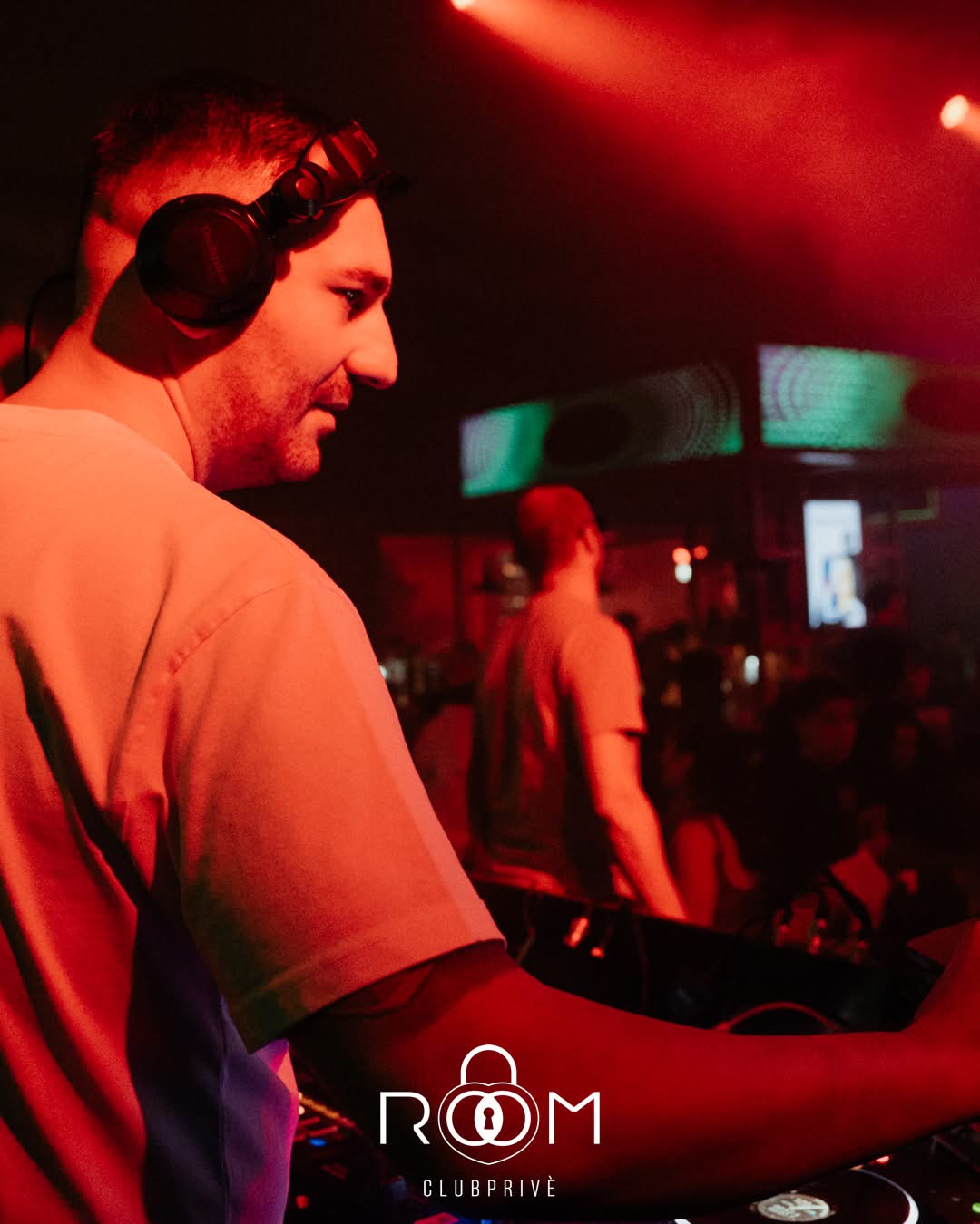 Il DJ con le cuffie in consolle, luce rossa e schermi verdi alle spalle.