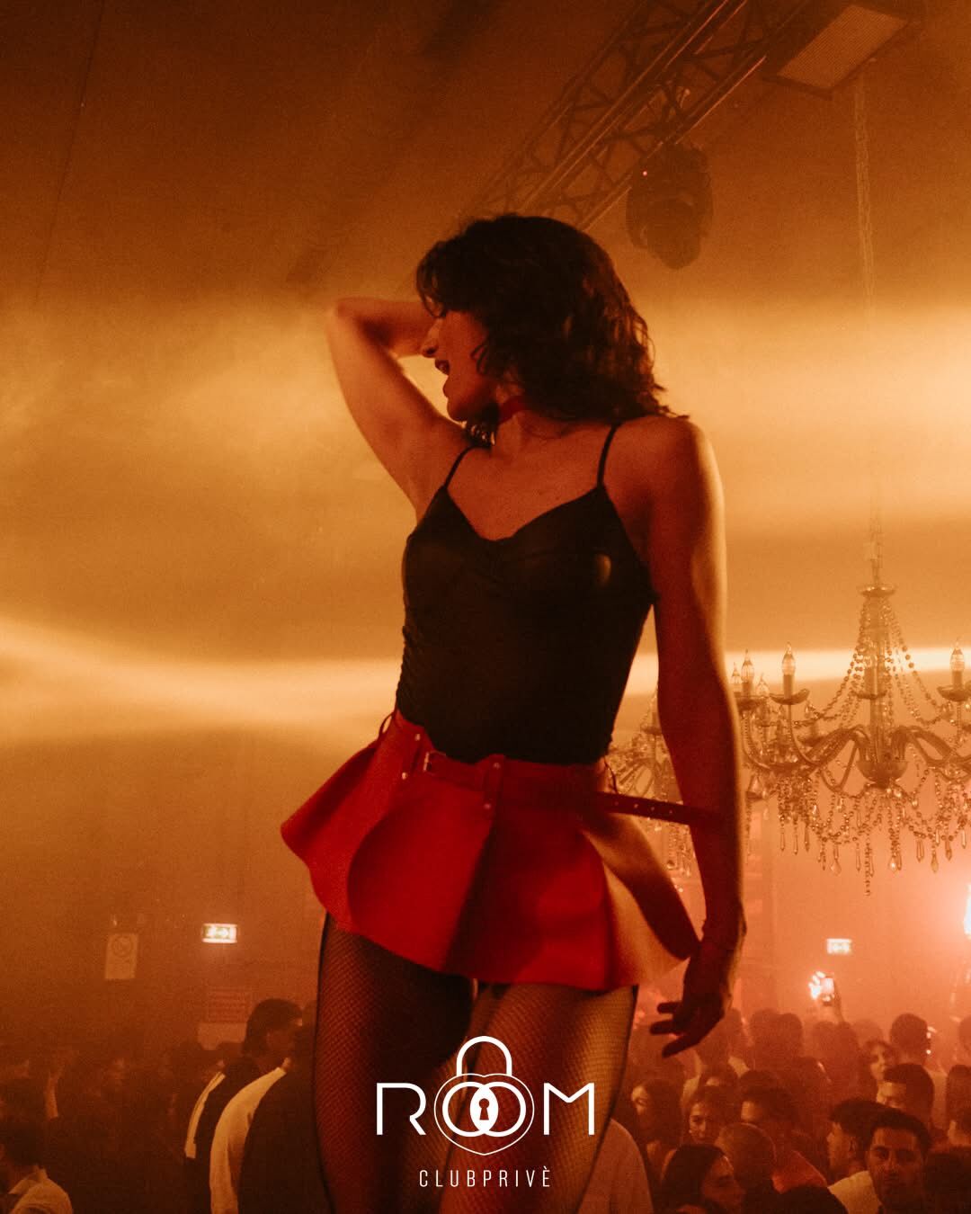 Ballerina in abito nero e minigonna rossa si esibisce sul palco tra i fasci di luce e i lampadari del locale.
